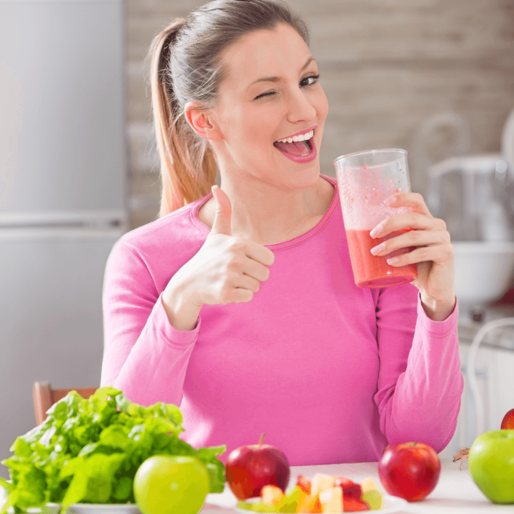 une femme fait un clin œil et souri devant des légumes en tenant un verre de jus de fruits